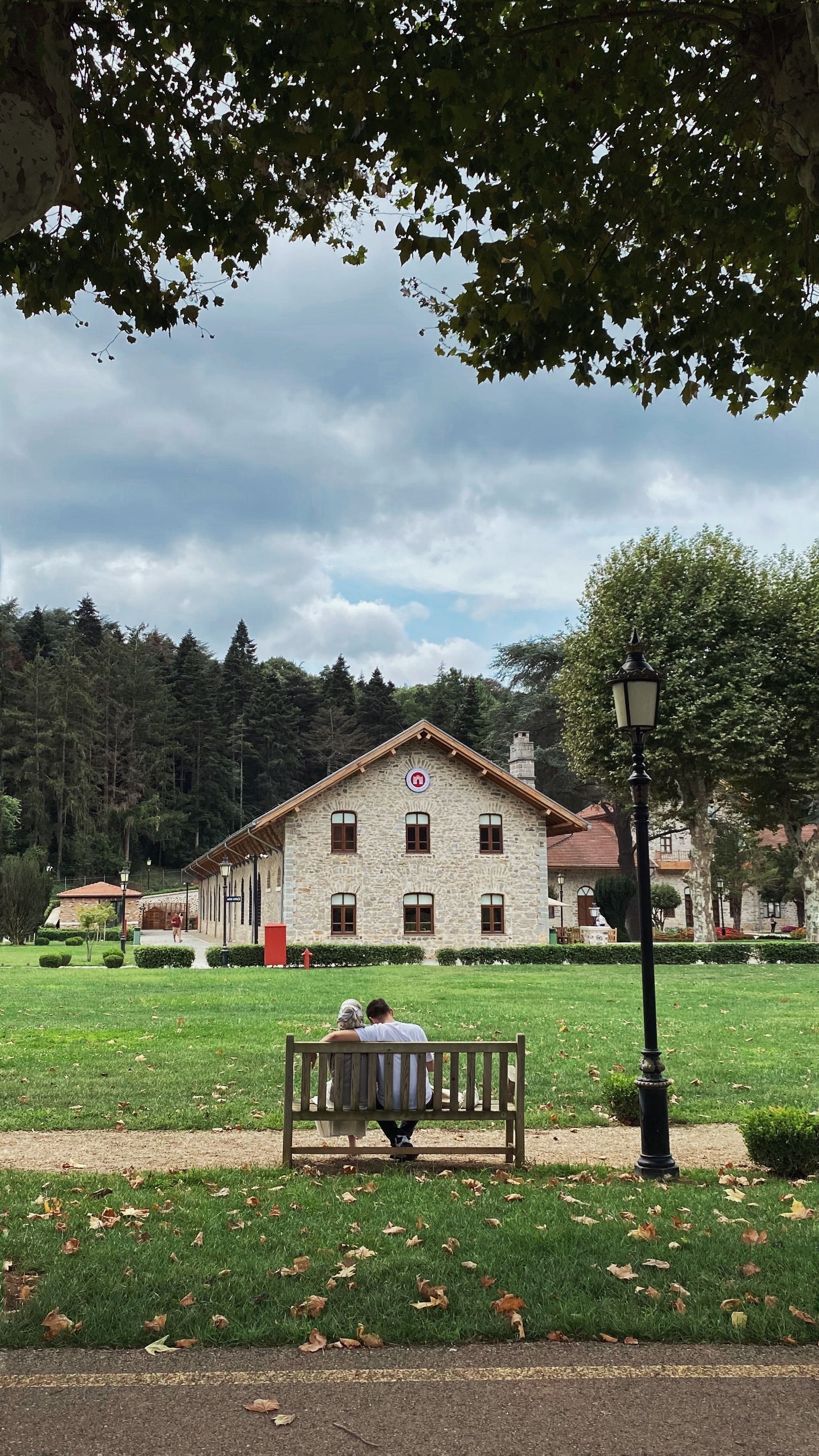 A couple sits on a park bench facing a quaint stone building amidst lush greenery.