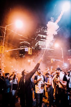 Vibrant city celebration at night with smoke flares and crowds gathered under illuminated skyscrapers.