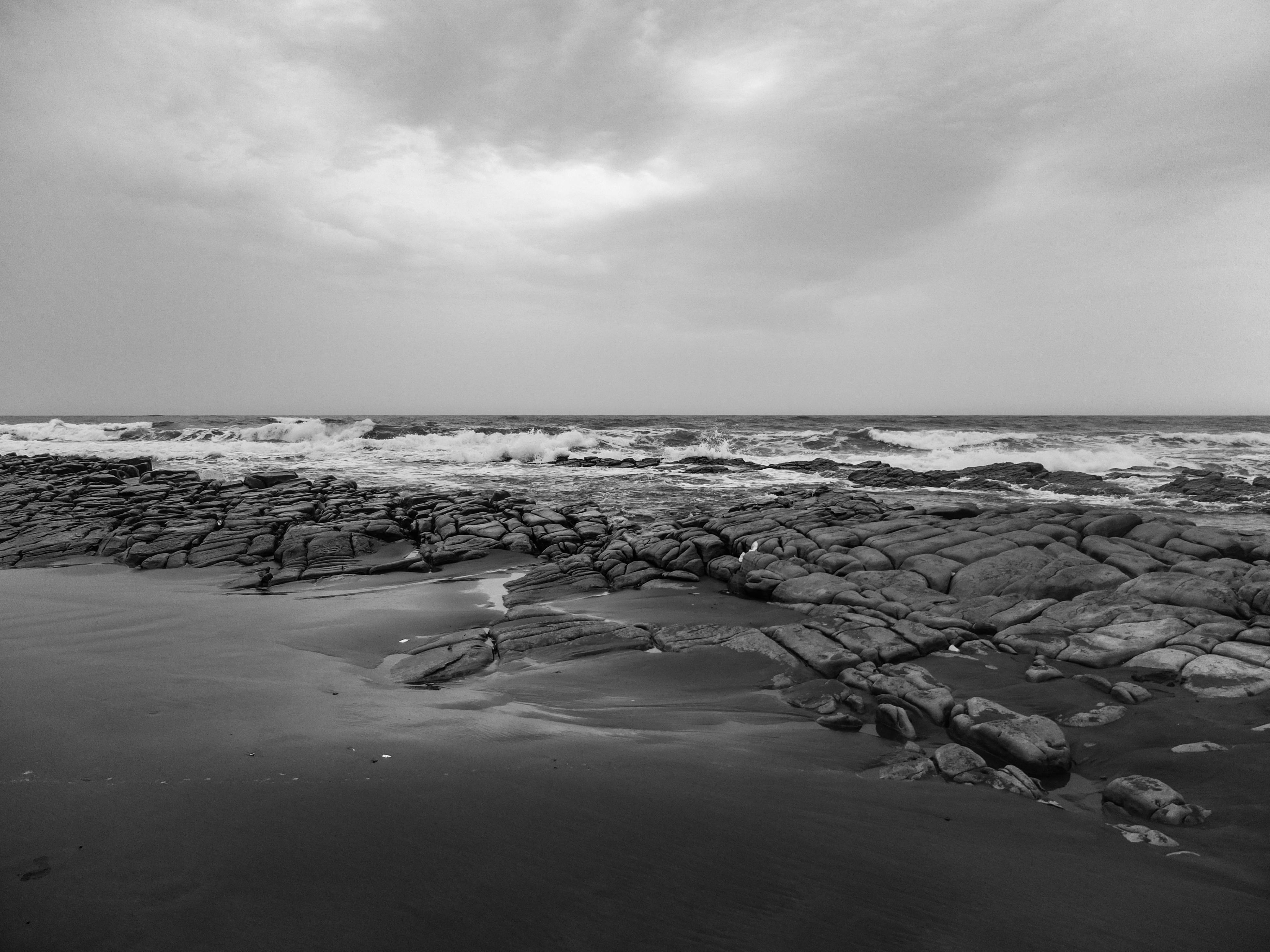Monochrome view of waves crashing on rocky coast under cloudy sky.