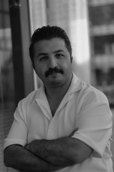 Elegant black and white portrait of a man with a mustache, arms crossed, wearing a white shirt, in an urban setting.