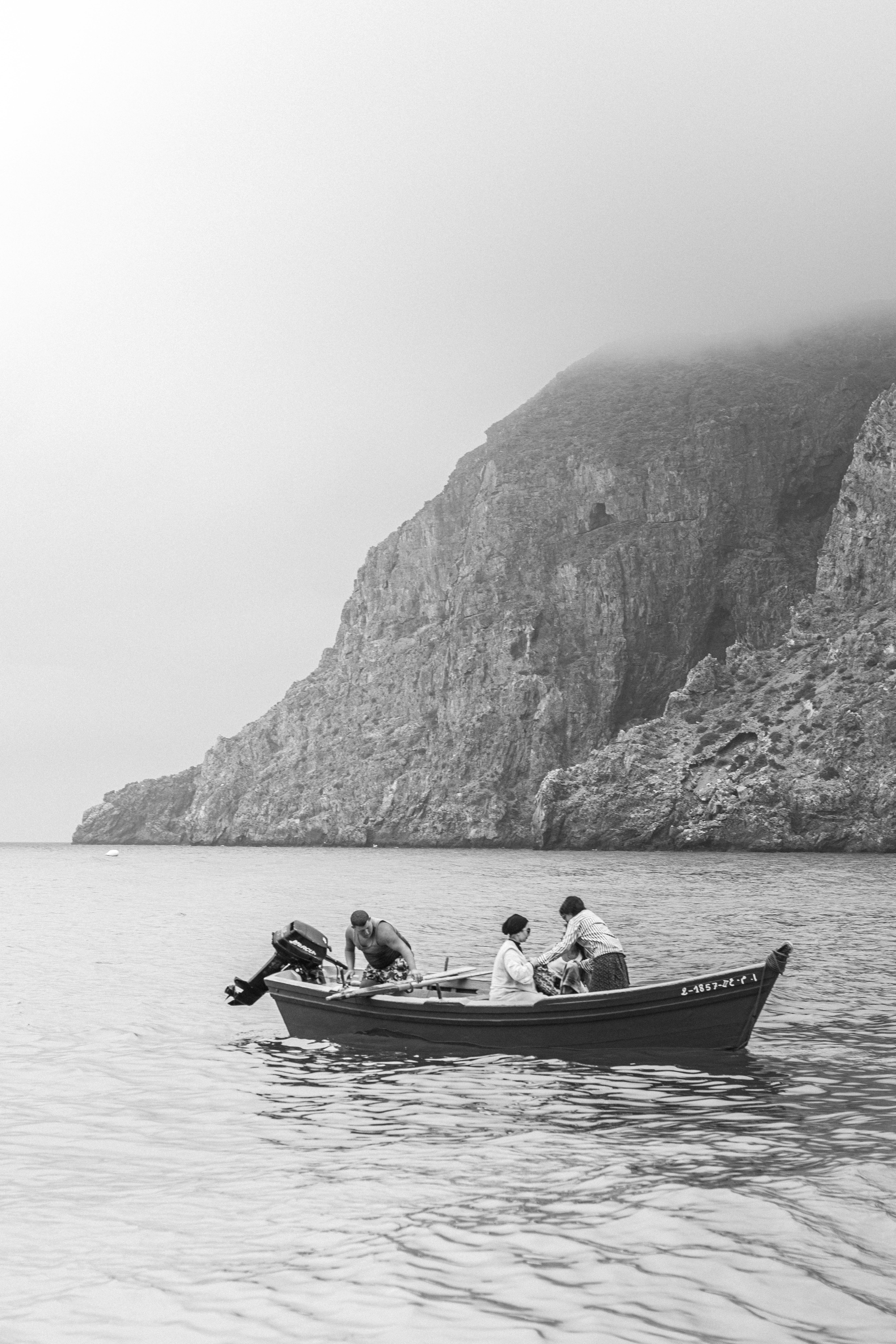 Black and white image of a motorboat at sea with people near a misty rocky coastline.