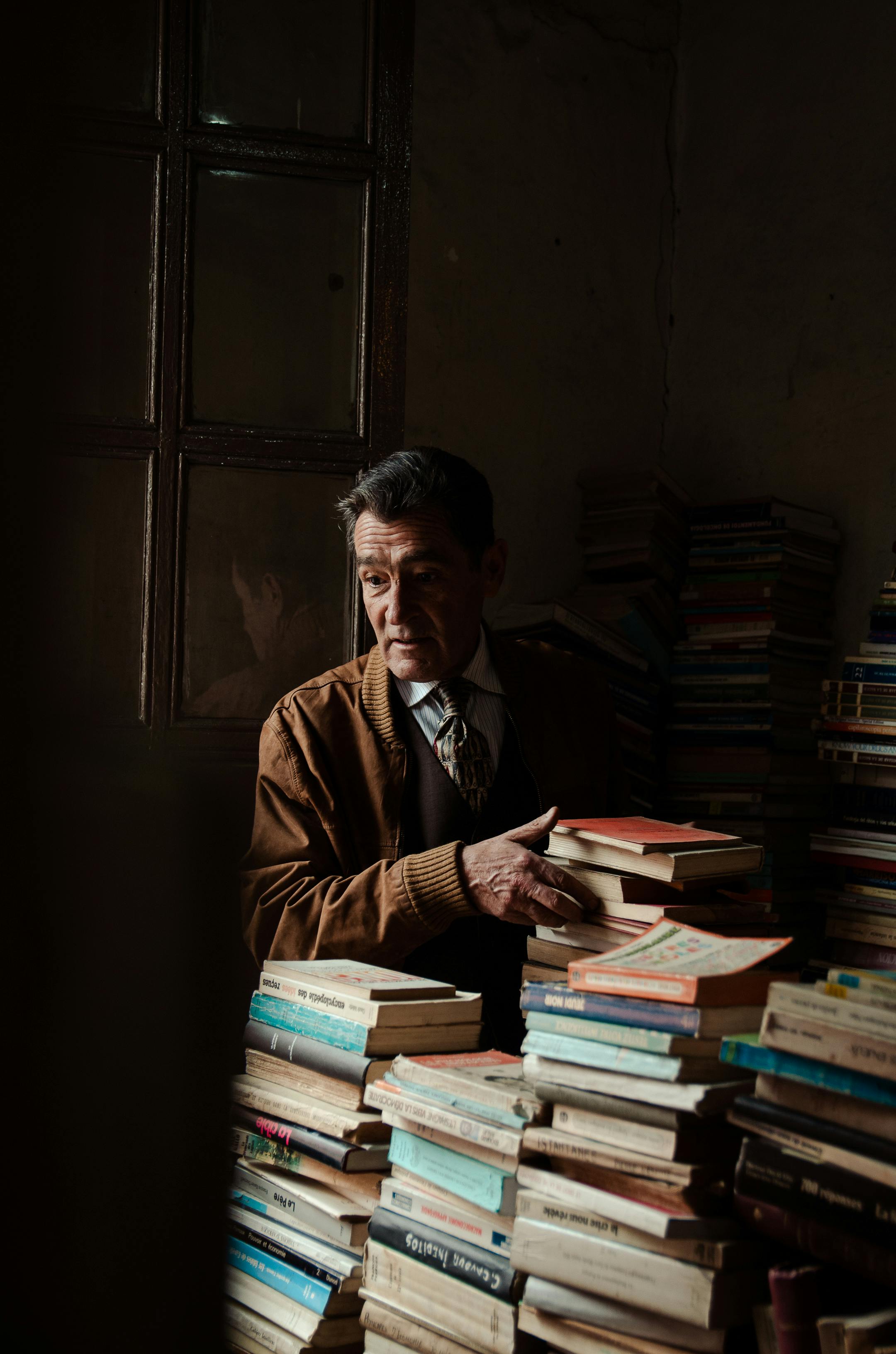 An elderly man surrounded by tall stacks of books in a cozy vintage bookstore.