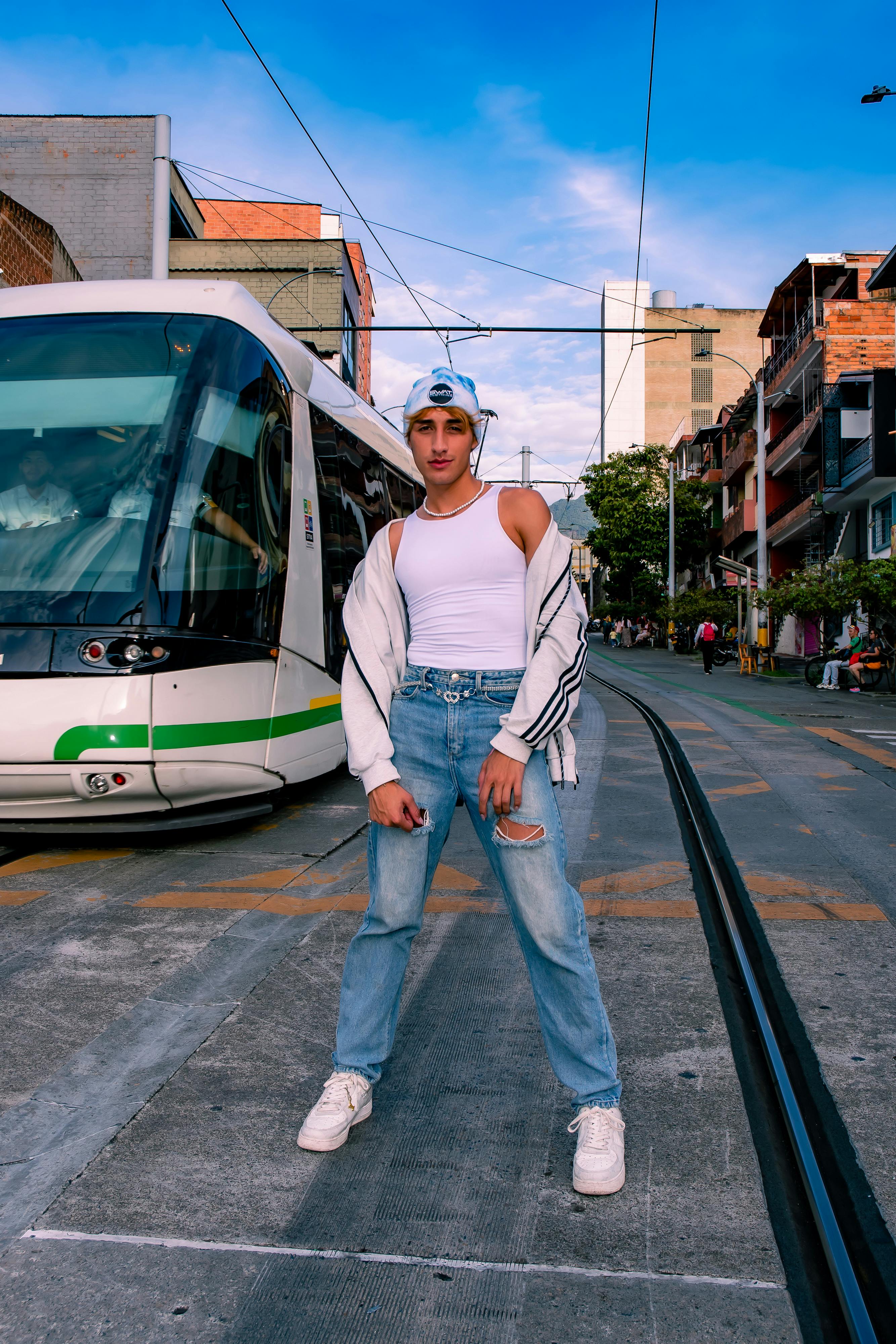 A young adult in stylish attire poses confidently by a tram in Medellín, Colombia's vibrant cityscape.