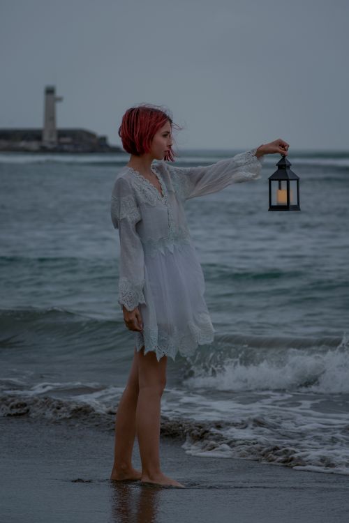 Free A woman with red hair holds a lantern at the seashore near a lighthouse, creating a dramatic seaside scene. Stock Photo