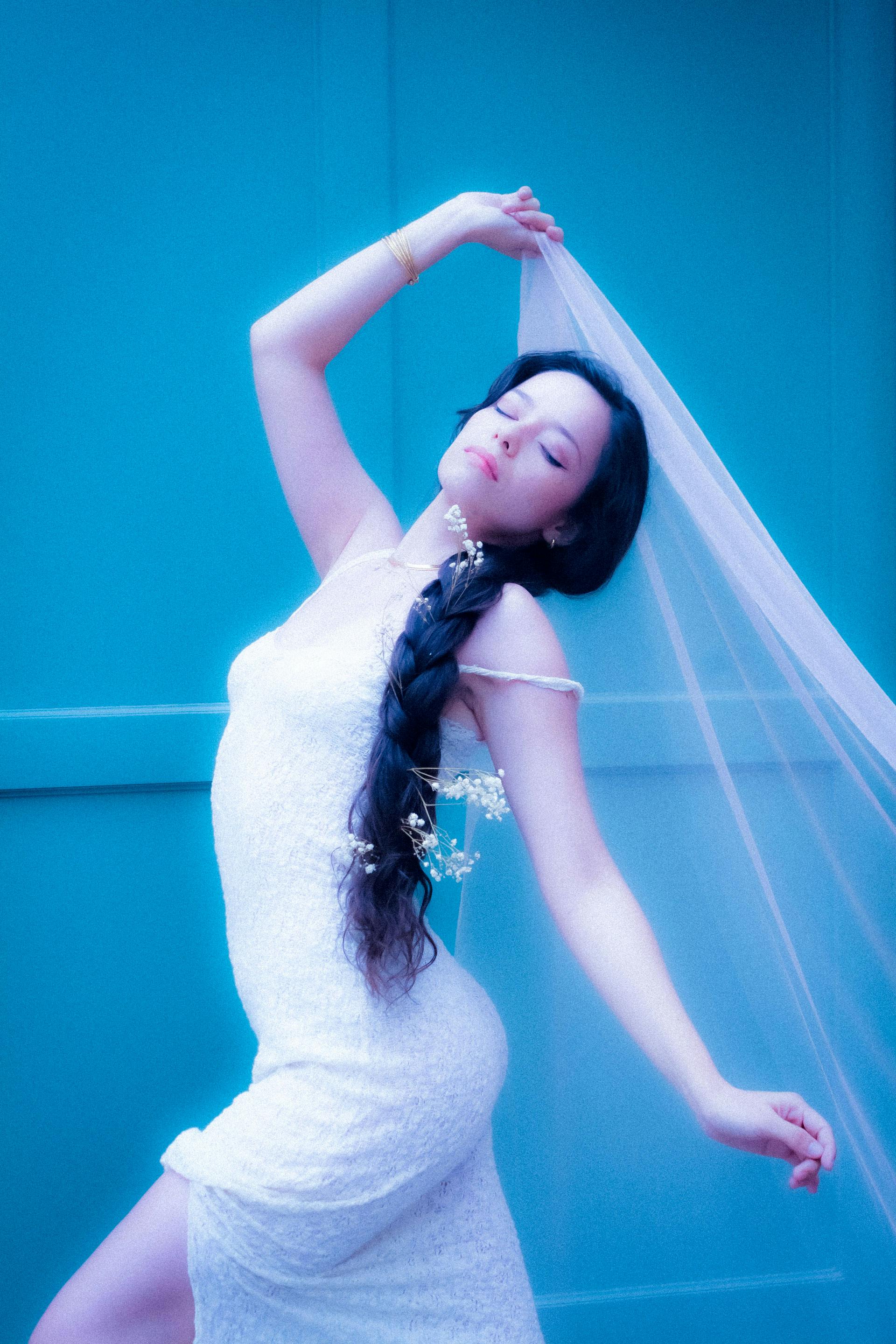 A graceful bride in a white dress poses elegantly with a veil in a vibrant blue setting.