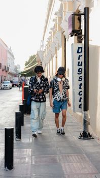 Two young adults walk along a sunny city sidewalk, passing by a drugstore sign.