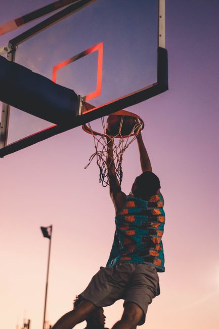 A basketball player successfully dunks as the sun sets, creating a dramatic silhouette.