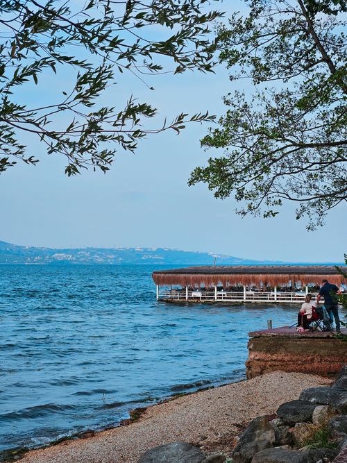 Free A peaceful lakeside scene with a wooden jetty and people enjoying a picnic under trees. Stock Photo
