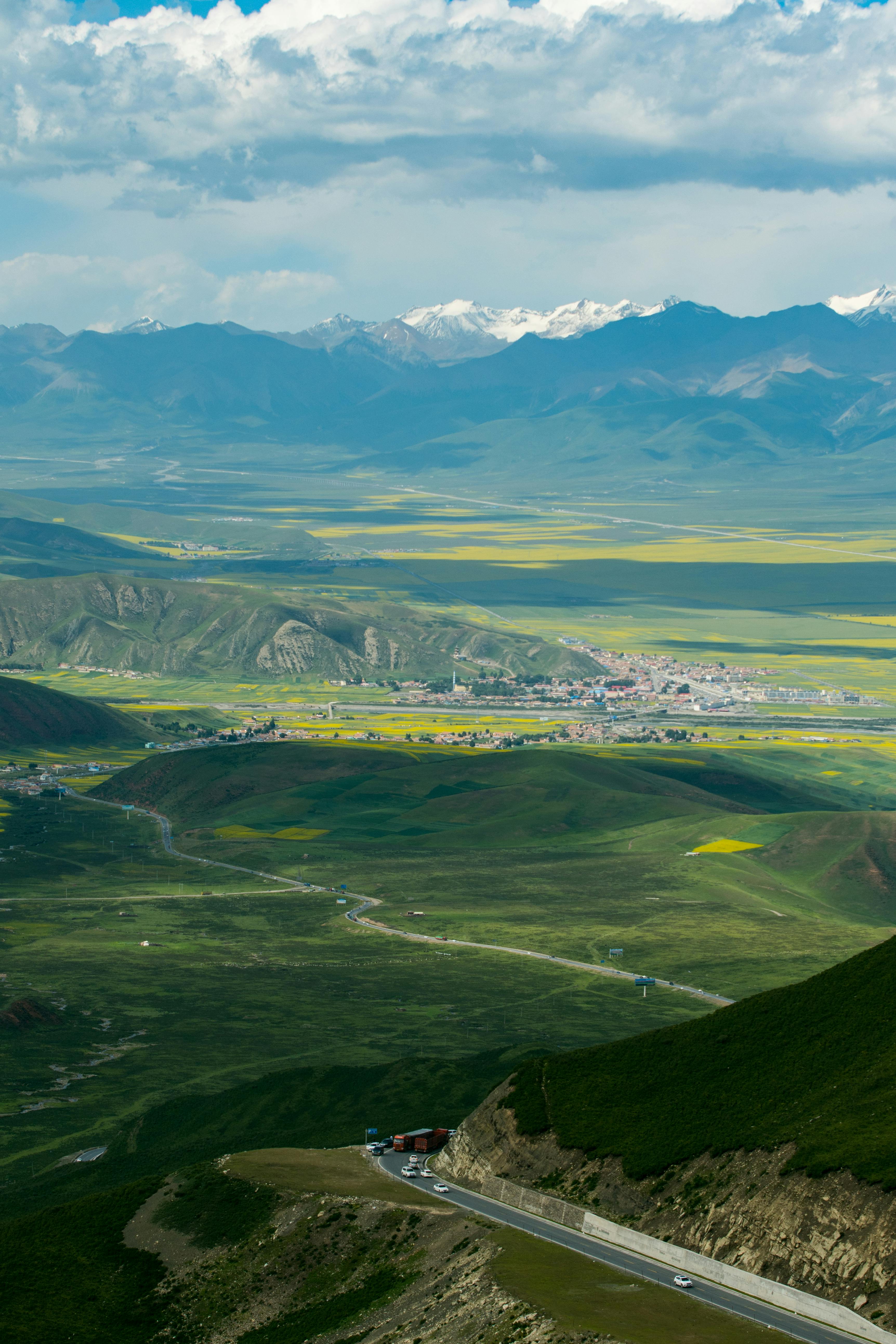 Panoramic view of a lush valley with mountains, a winding road, and a distant village under blue skies.