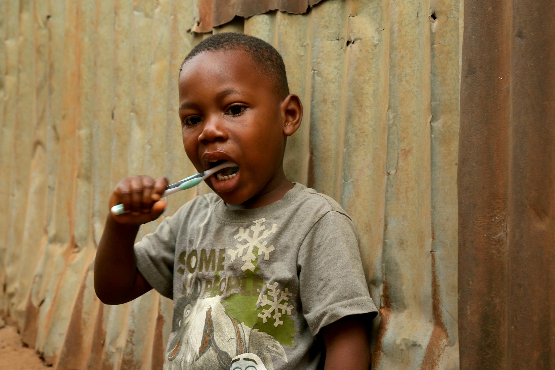 Young boy practicing oral hygiene outdoors near a corrugated metal wall in Freetown, Sierra Leone.