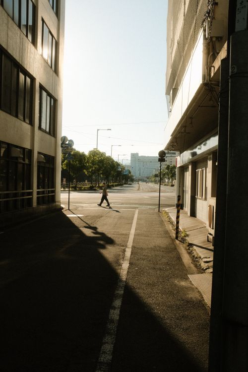 Free A pedestrian crosses an urban street with sunlight casting shadows between buildings. Stock Photo