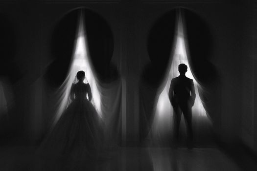 Artistic black and white silhouettes of a bride and groom by window curtains, creating a dramatic wedding atmosphere.