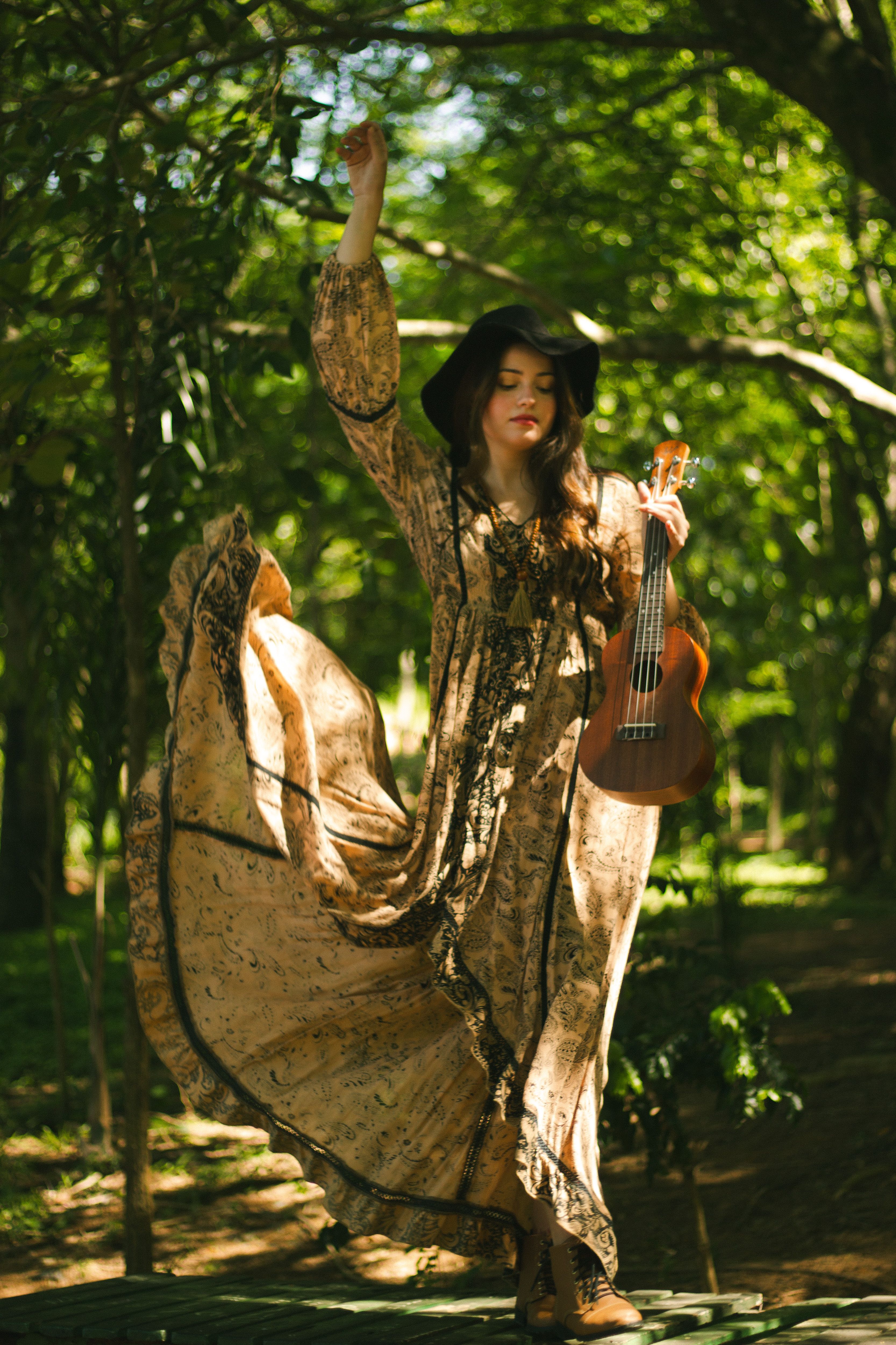 A bohemian-style woman plays a ukulele in a sunlit forest, surrounded by lush greenery.
