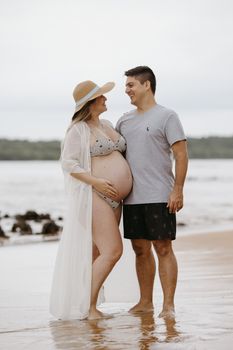 A smiling pregnant couple stands together on a serene beach, enjoying the coastal ambiance.