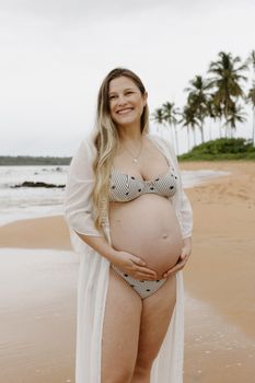 A cheerful pregnant woman stands on a tropical beach, embracing motherhood joyfully.