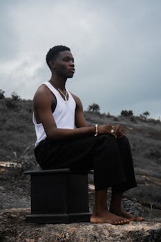 A contemplative young man sits outdoors on a rock, stylishly dressed in black pants and a sleeveless top.
