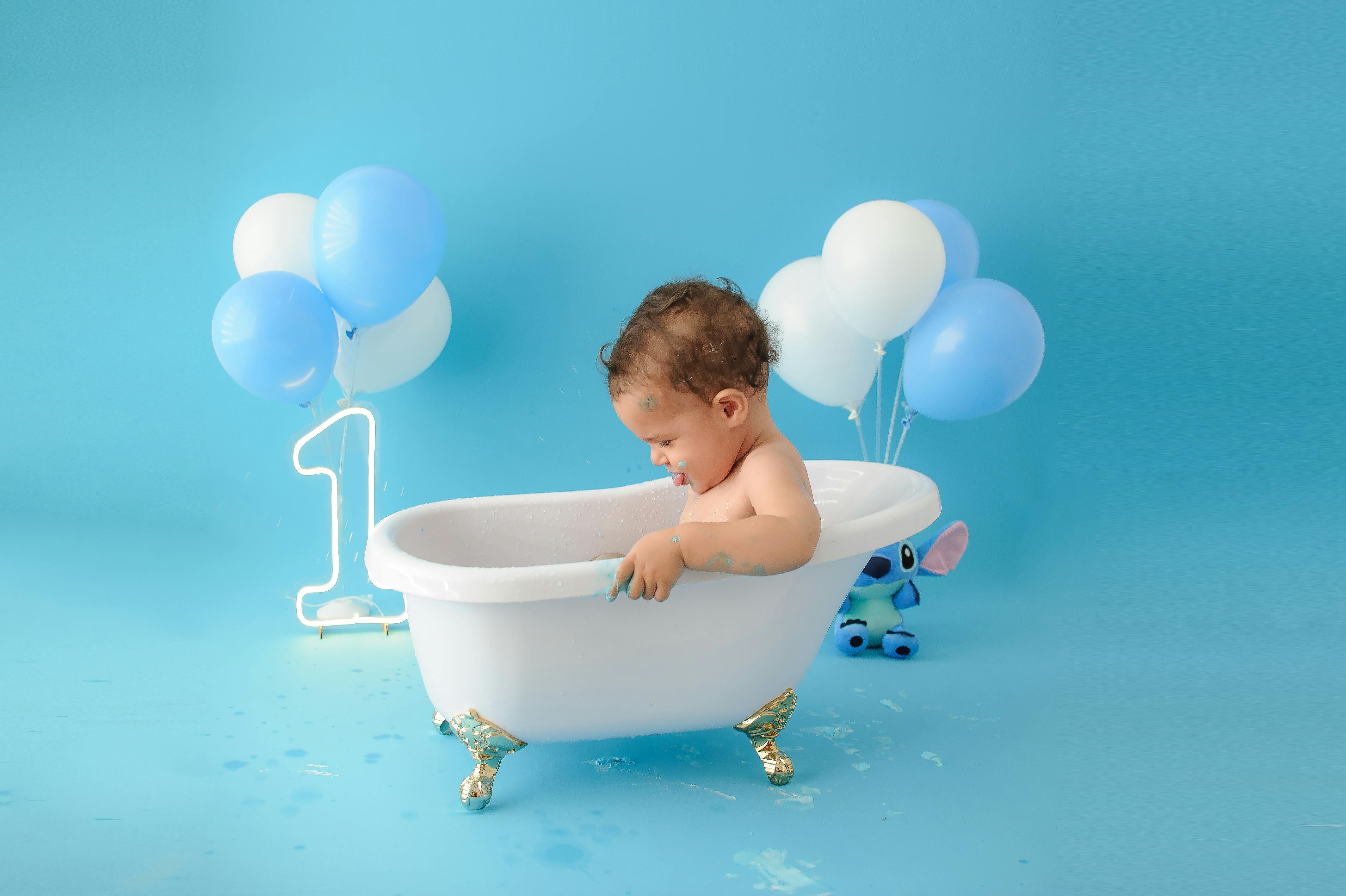 Adorable baby enjoying a bath in a white tub with balloons. Perfect for first birthday themes.