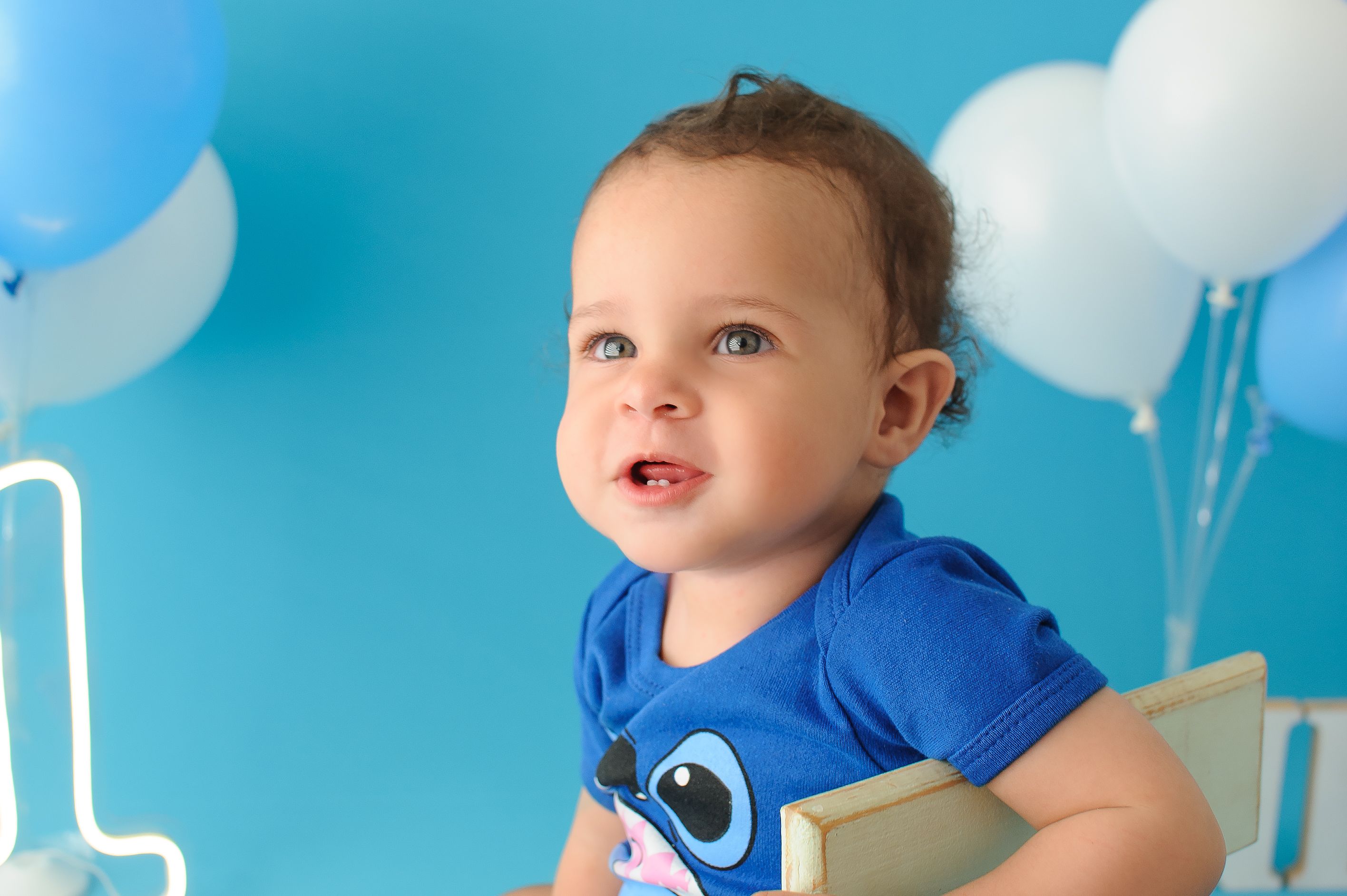 Cheerful baby in blue shirt enjoying first birthday with balloons in a studio shoot. | BocaVibe