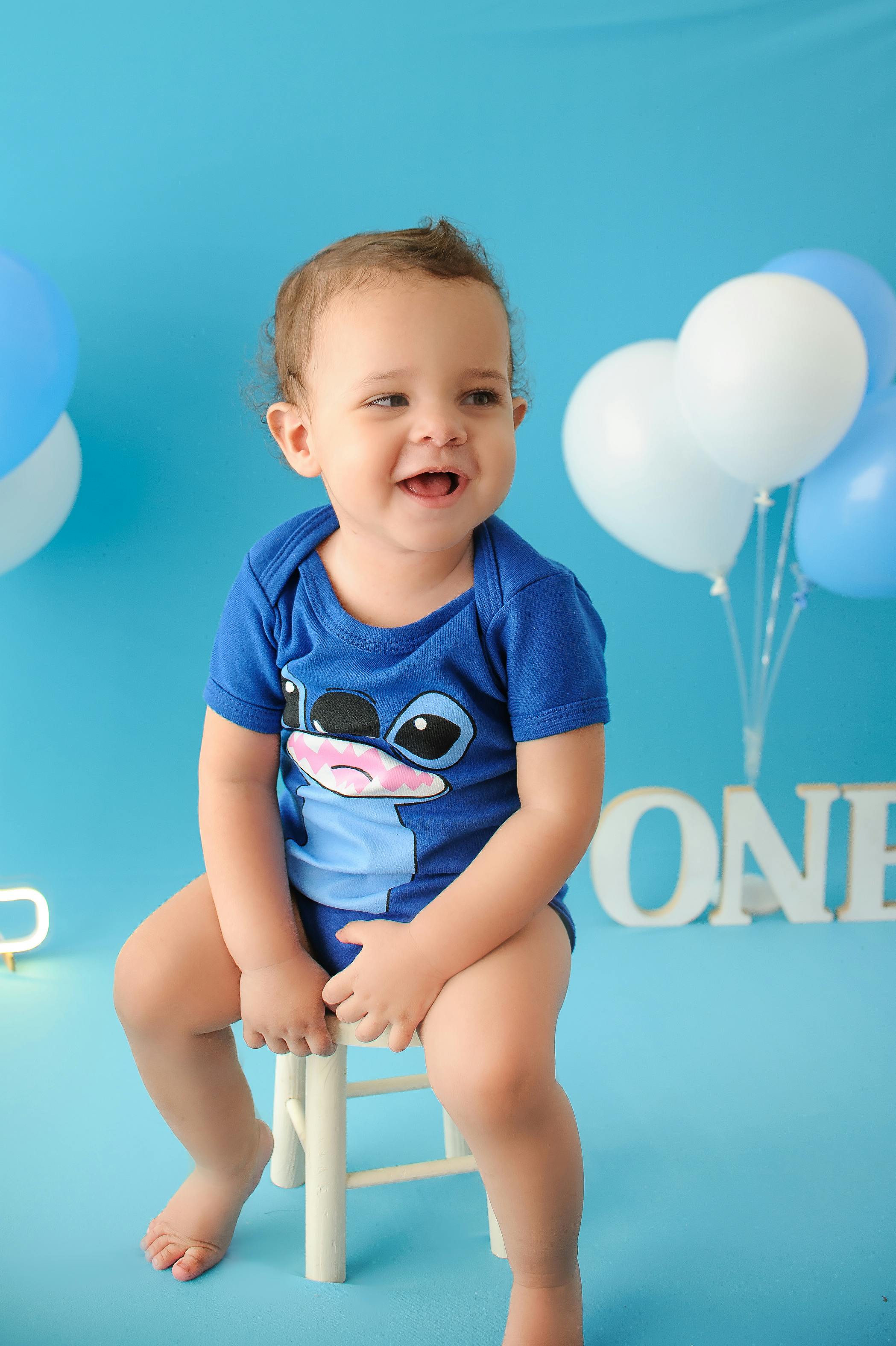 Smiling baby boy on chair with balloons, enjoying a studio shoot for his first birthday.