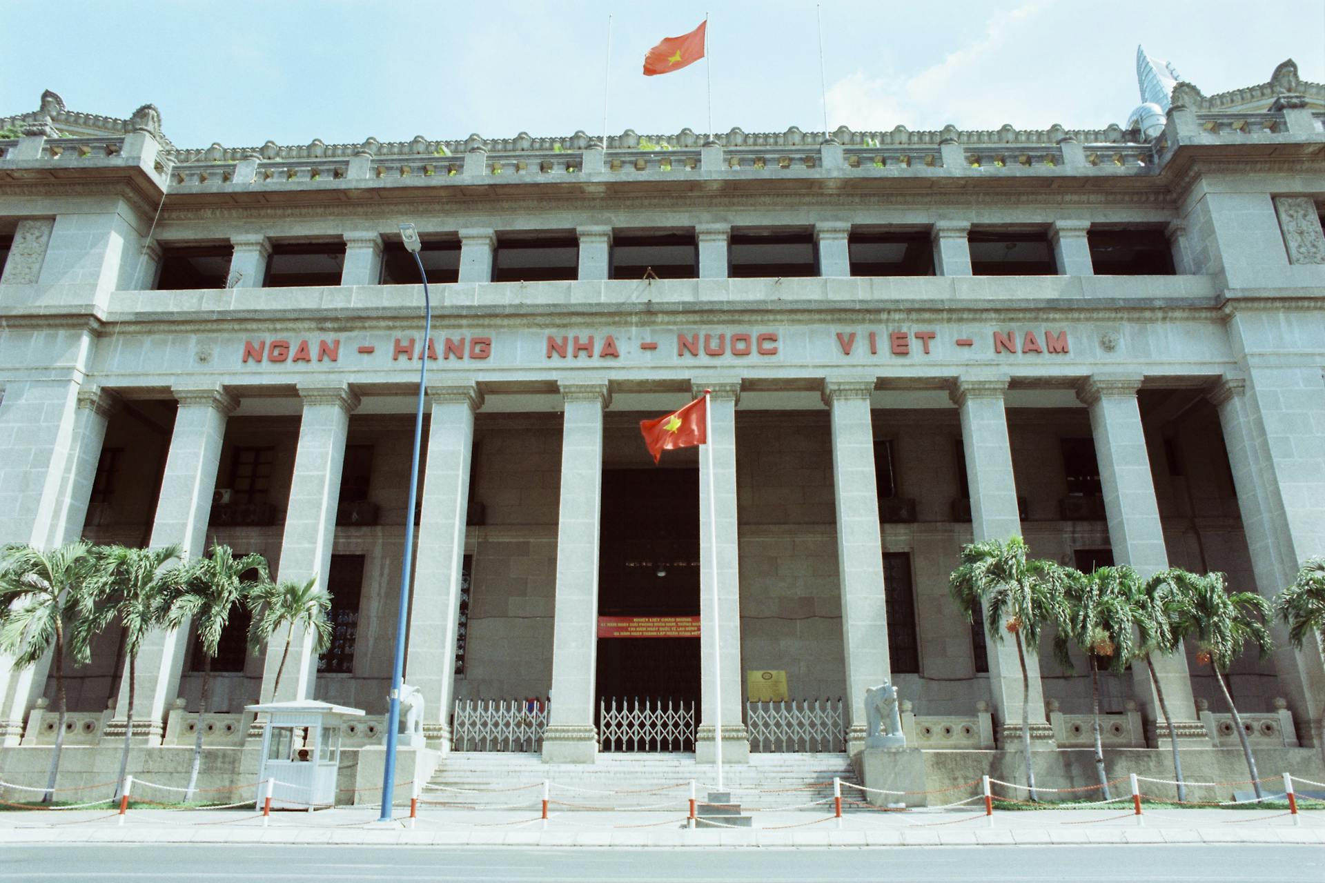 Facade of the State Bank of Vietnam with palm trees and Vietnamese flags.