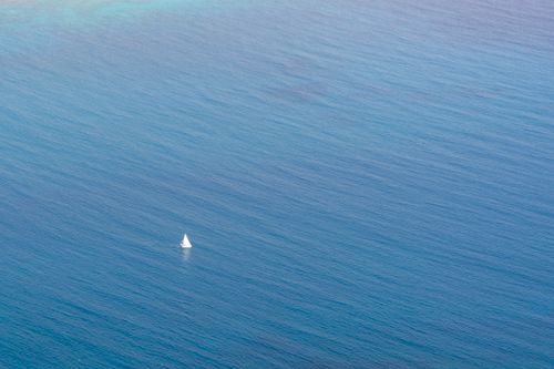 Free A small sailboat sailing across a vast, tranquil blue ocean, viewed from above. Stock Photo