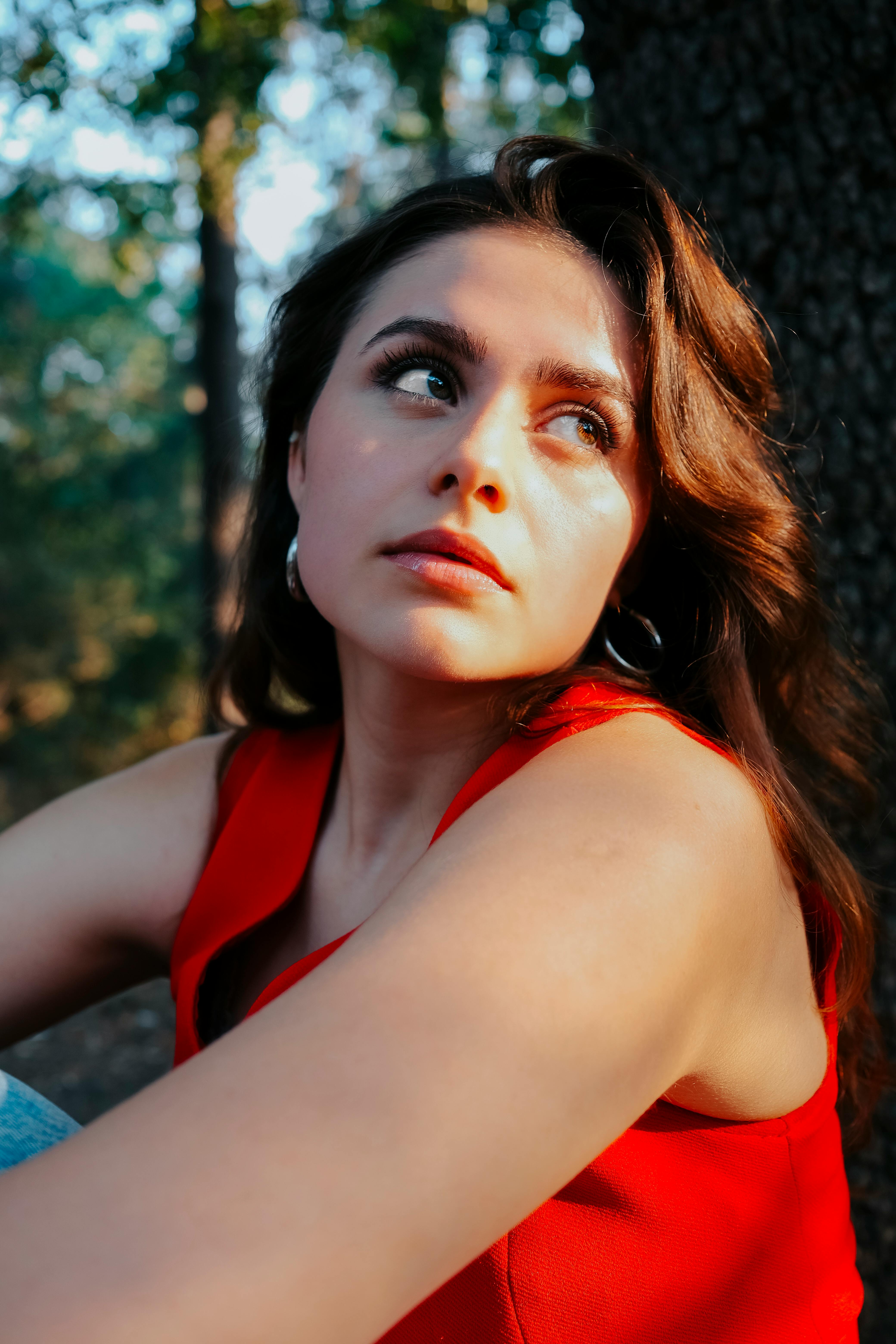 A contemplative woman in a red top sitting against a tree in a sunlit forest setting.
