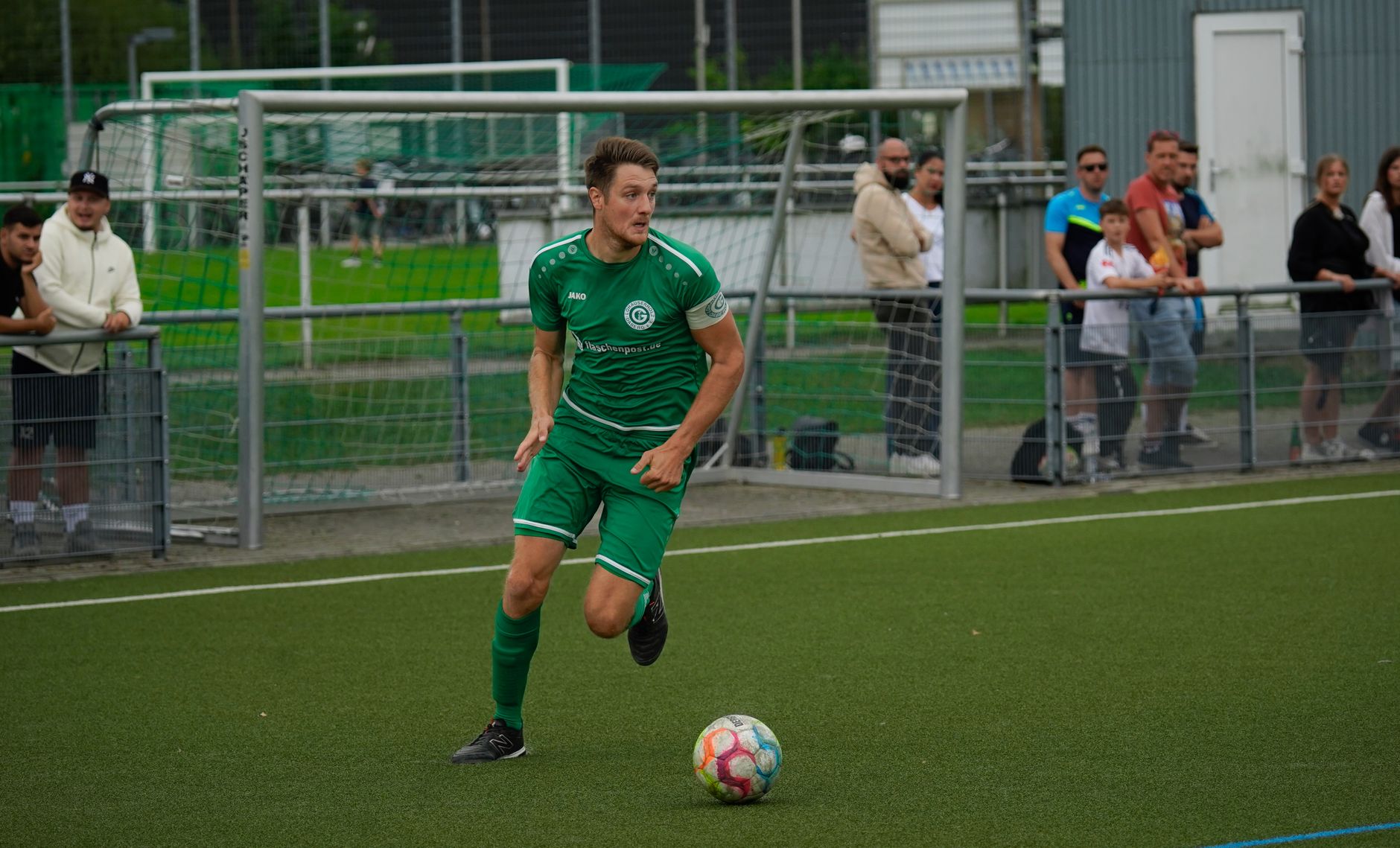 Adult male soccer player in green jersey dribbling ball on pitch during daytime match with spectators.