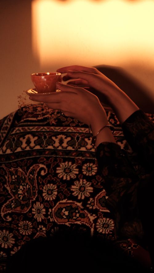 Free Artistic shot of hands holding a tea cup, illuminated by warm lighting over a patterned fabric. Stock Photo