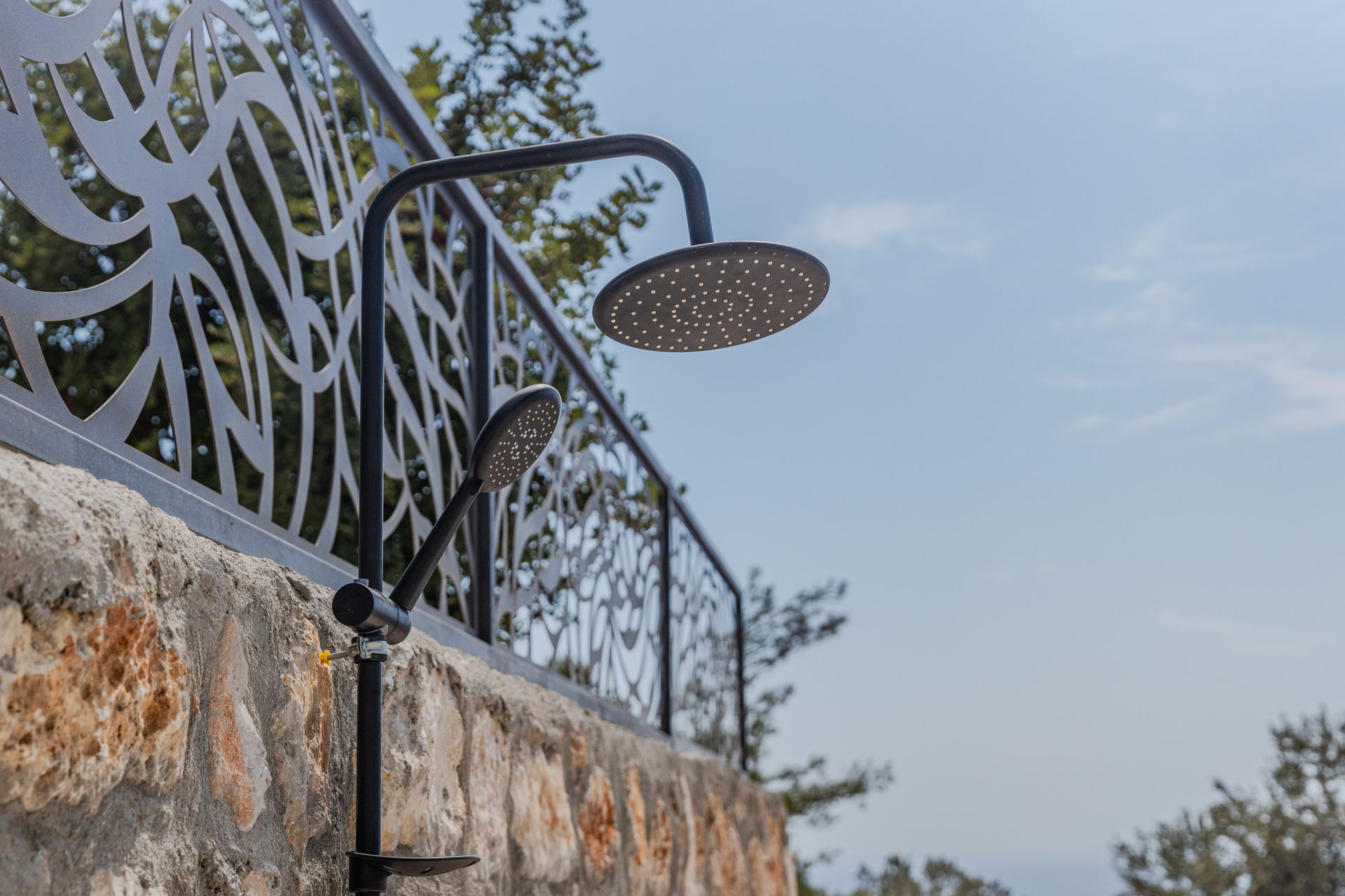 Outdoor shower setup with modern metal fence design against a stone wall under clear blue sky.