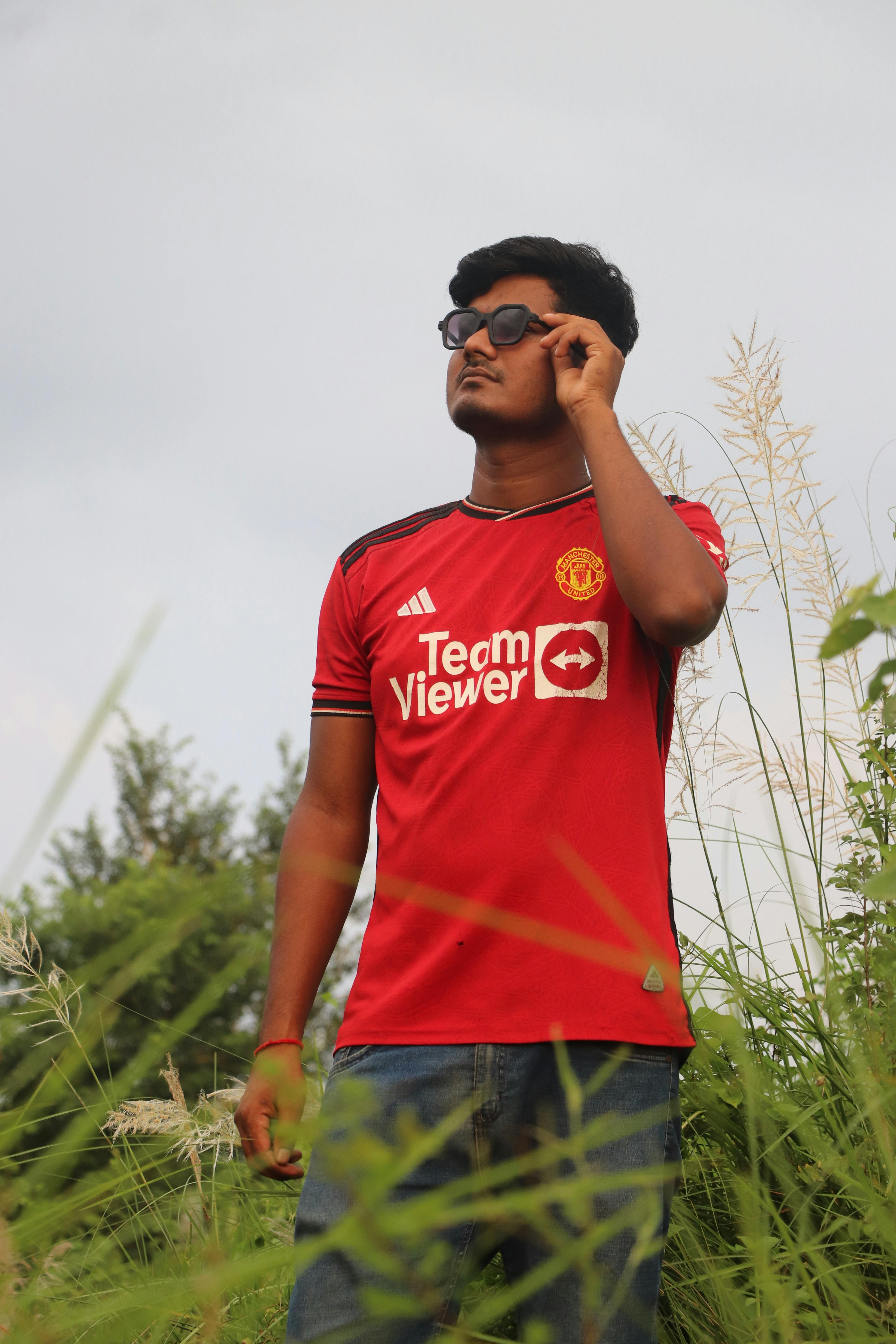 Young man wearing a red jersey adjusting sunglasses in an outdoor rural setting in Bangladesh.