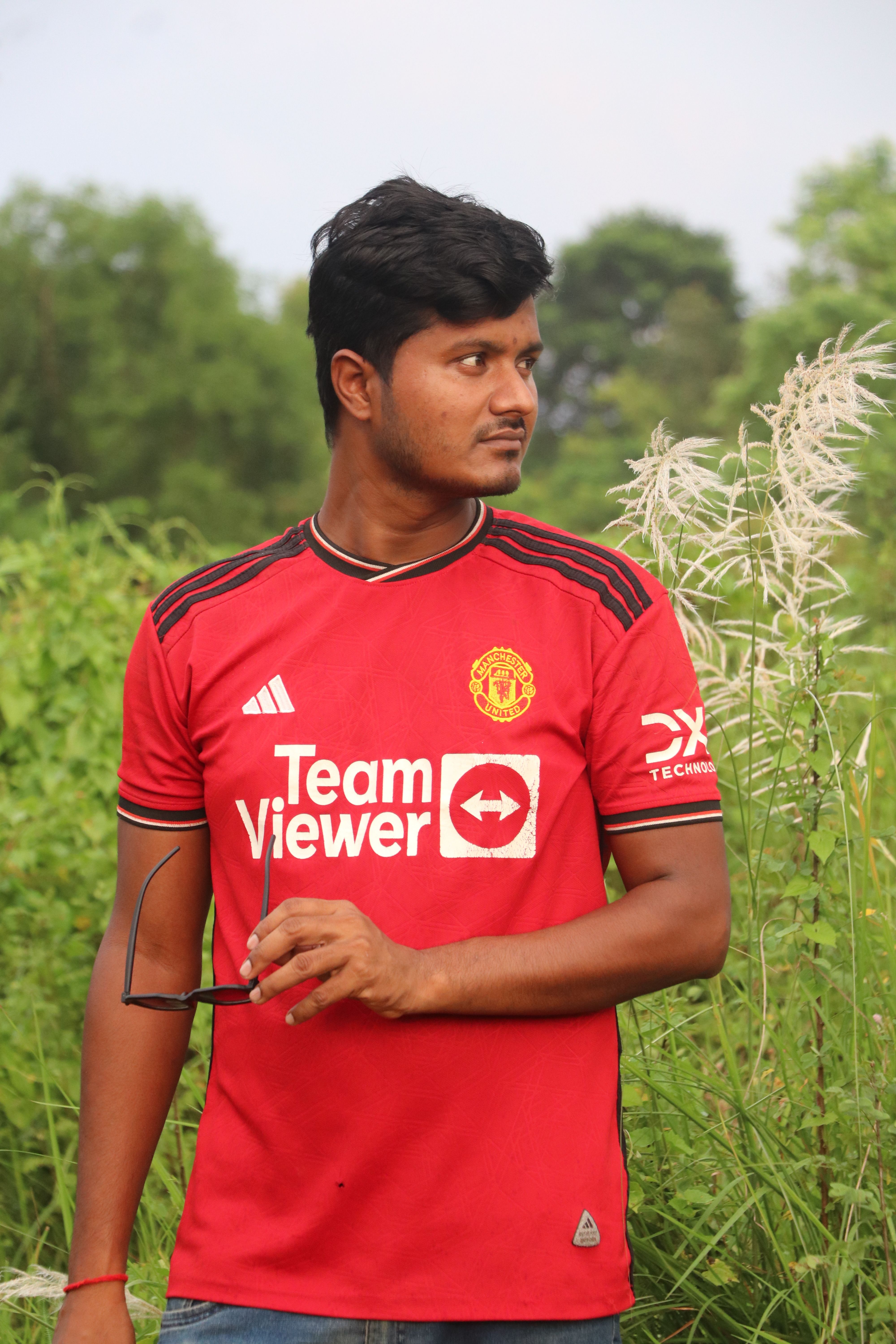 South Asian man standing in lush greenery wearing a sports shirt in Rangpur, Bangladesh.