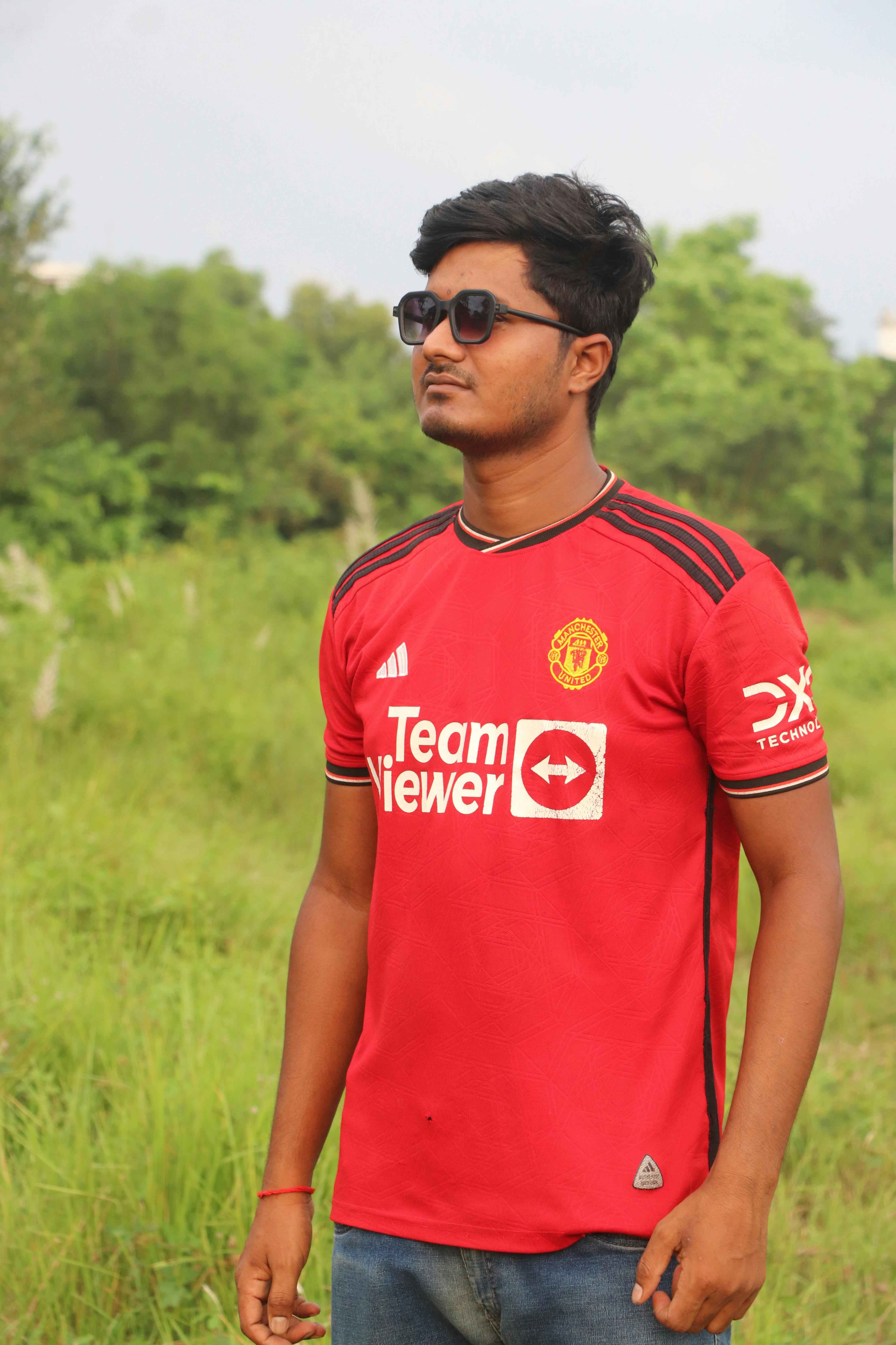 A young man in sunglasses standing outdoors in a red sports jersey, looking pensive.