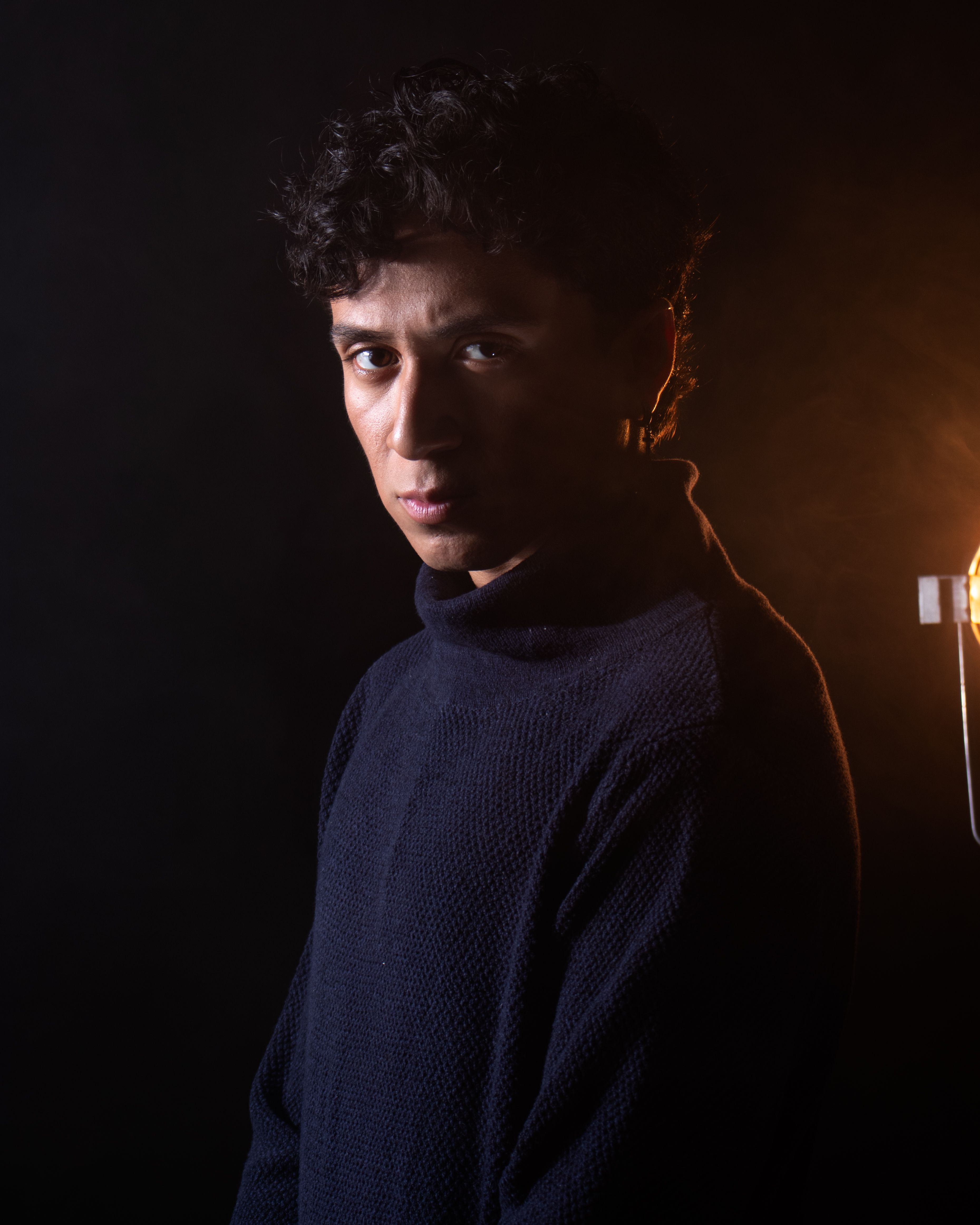 A striking portrait of a young man standing in a dark room, illuminated by warm dramatic lighting.