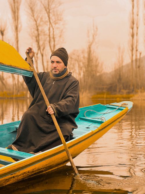 Free A man in a beanie paddles a colorful wooden boat on a tranquil lake during springtime. Stock Photo