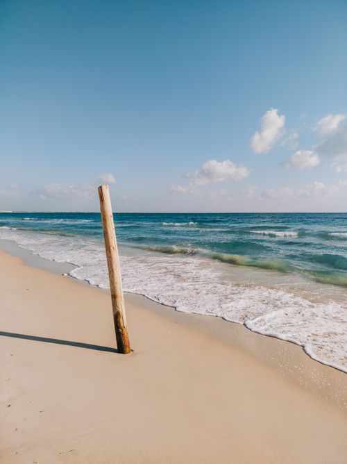 Free Serene beach scene featuring a wooden post on sandy shores with gentle waves under a clear blue sky. Stock Photo