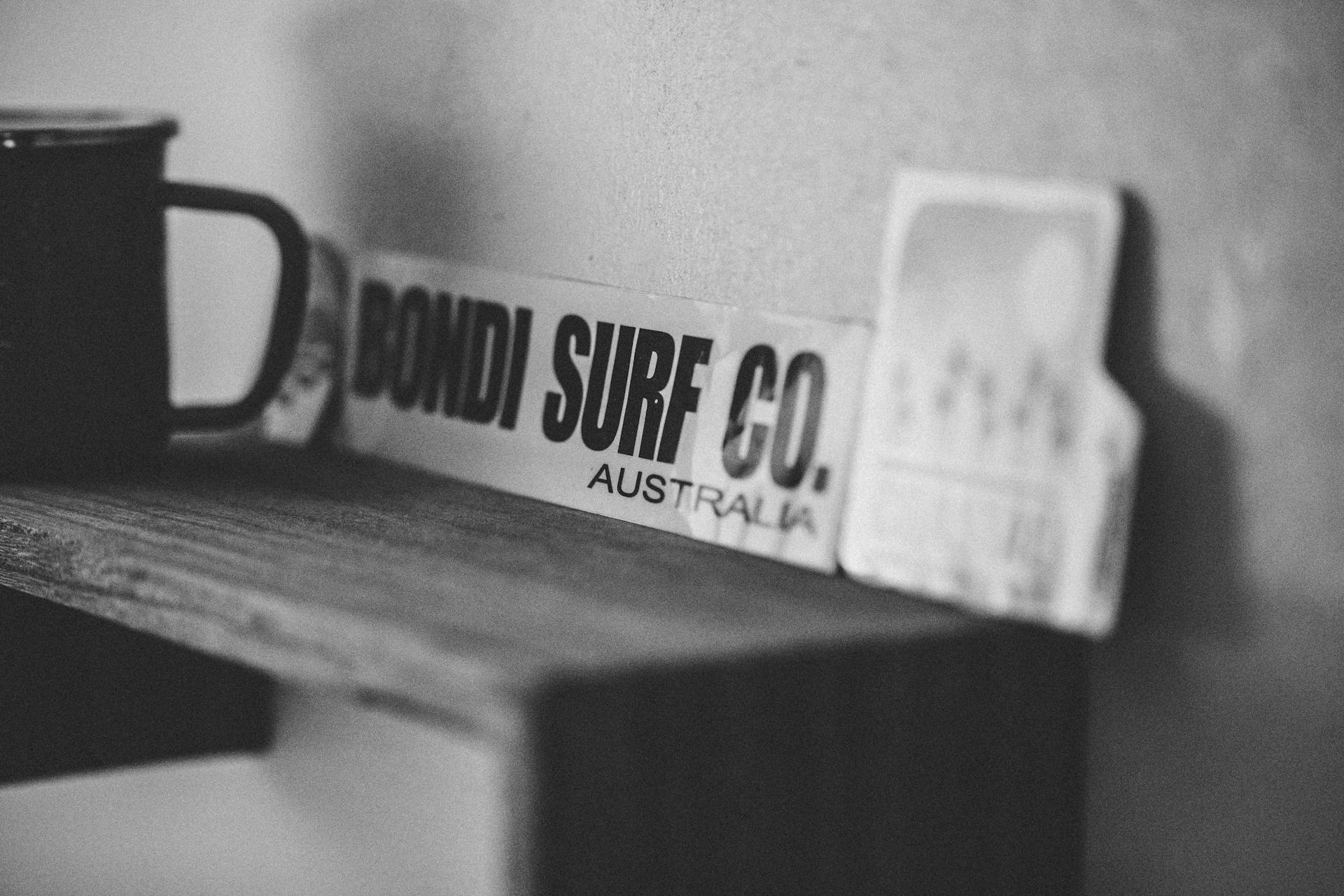 Black and white photo of a Bondi Surf Co board on a wooden shelf with a mug.