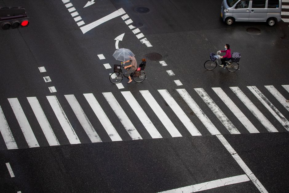 Cyclists navigate a rainy city crosswalk with umbrellas for protection.