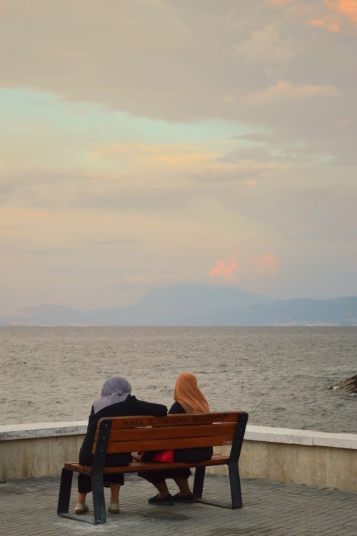 Free Two women in hijabs sitting on a bench by the sea during sunset. Stock Photo