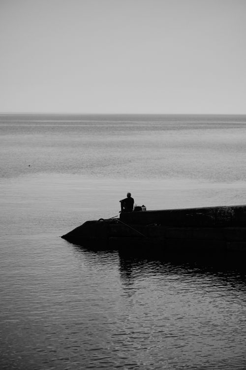 Free A lone person sitting on the dock, gazing at the tranquil sea in black and white. Stock Photo