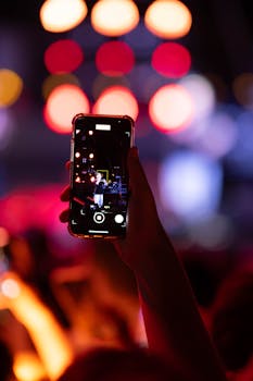 A hand holds a smartphone, filming a concert with vibrant stage lights.