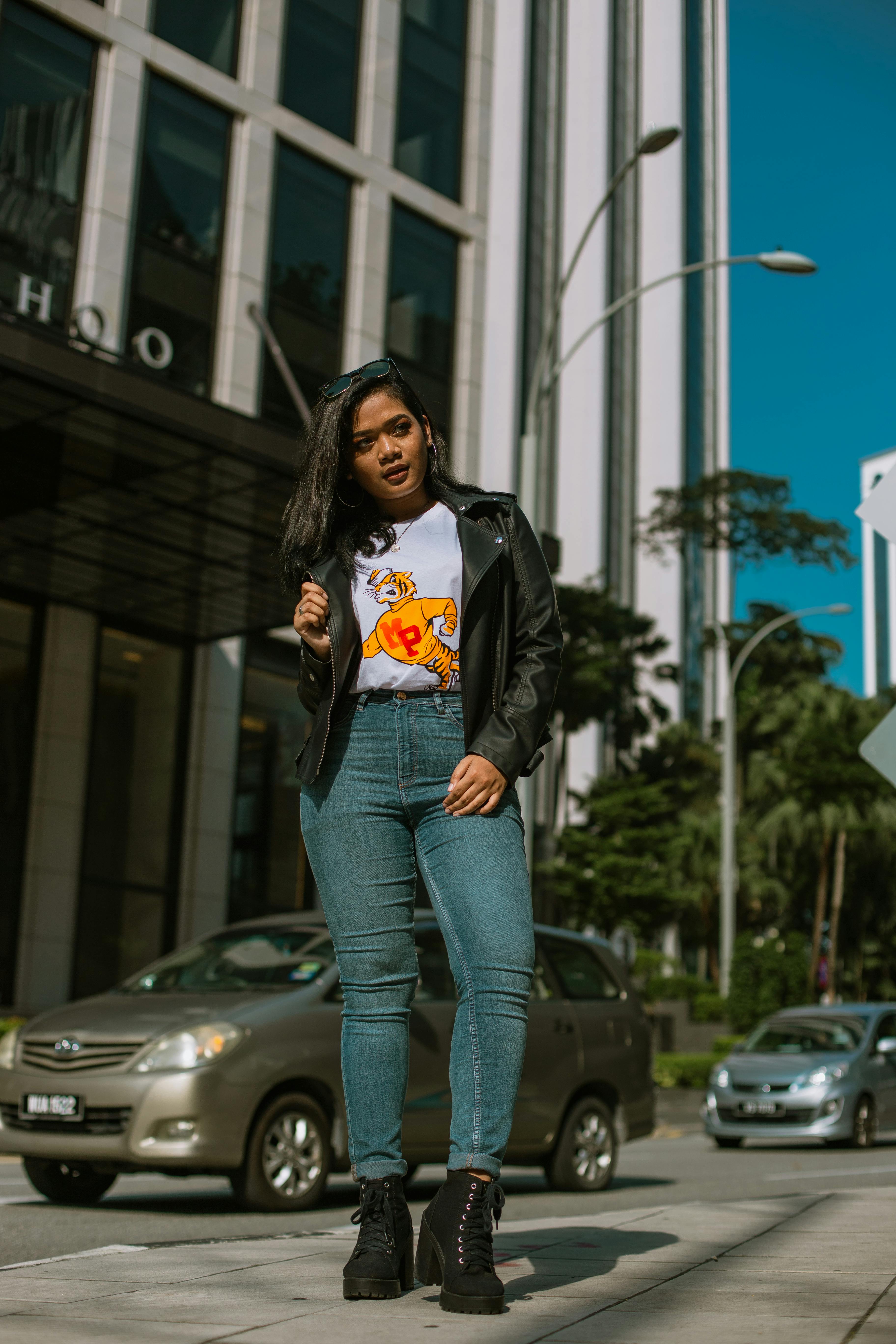 一位自信的女人身着时尚皮夹克和牛仔裤，在城市街道上摆出姿势，周围是来往的汽车