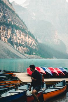 Man near colorful canoes on a dock with Rocky Mountains backdrop at Moraine Lake, Banff.