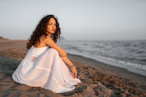 Wanita Mengenakan Gaun Tanpa Lengan Putih Duduk Di Pantai Dekat Perairan