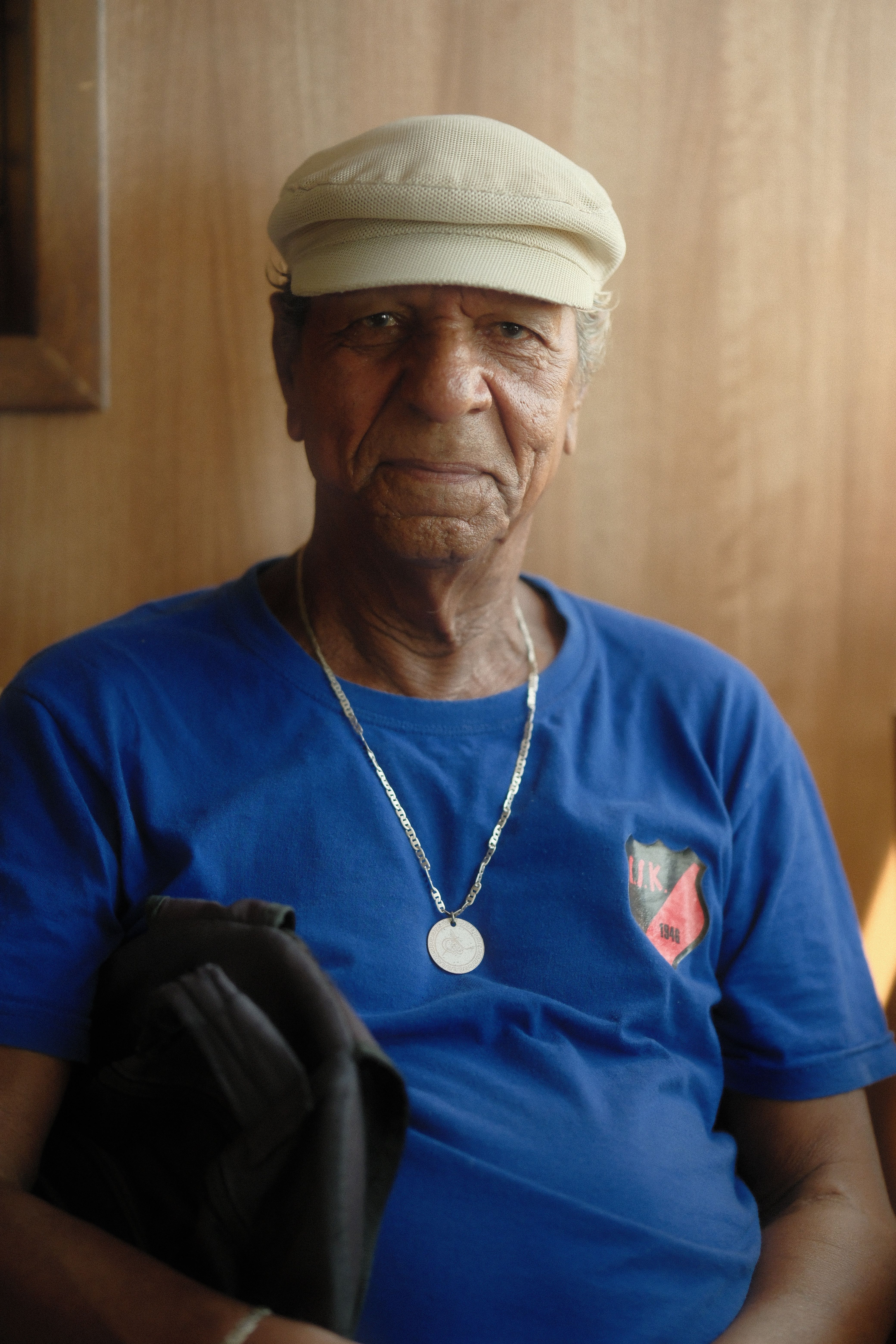 Elderly man in blue shirt and hat, sitting indoors with a gentle expression.