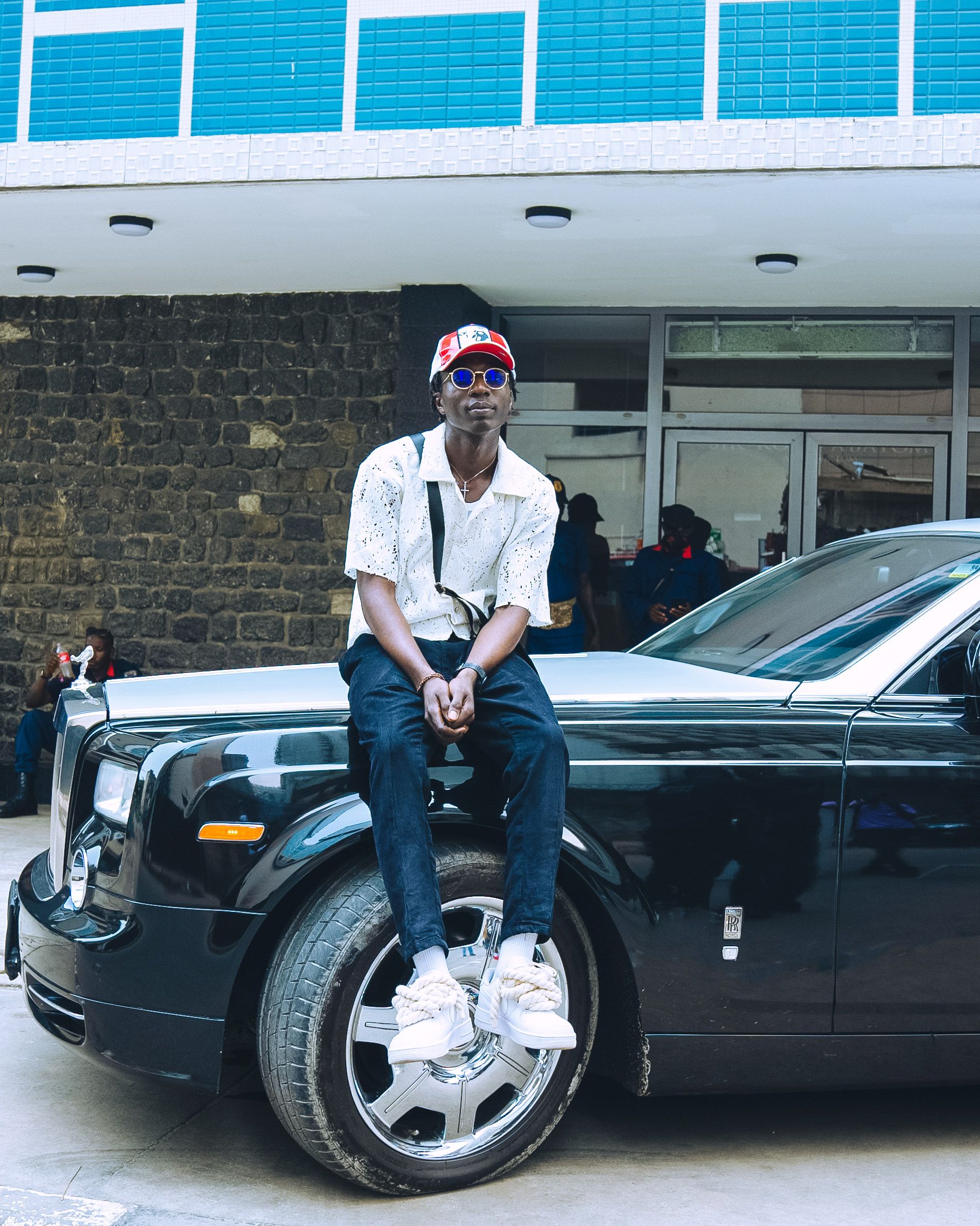 A fashionable man in sunglasses and cap sitting on a luxury car in an urban backdrop, exuding style and confidence.