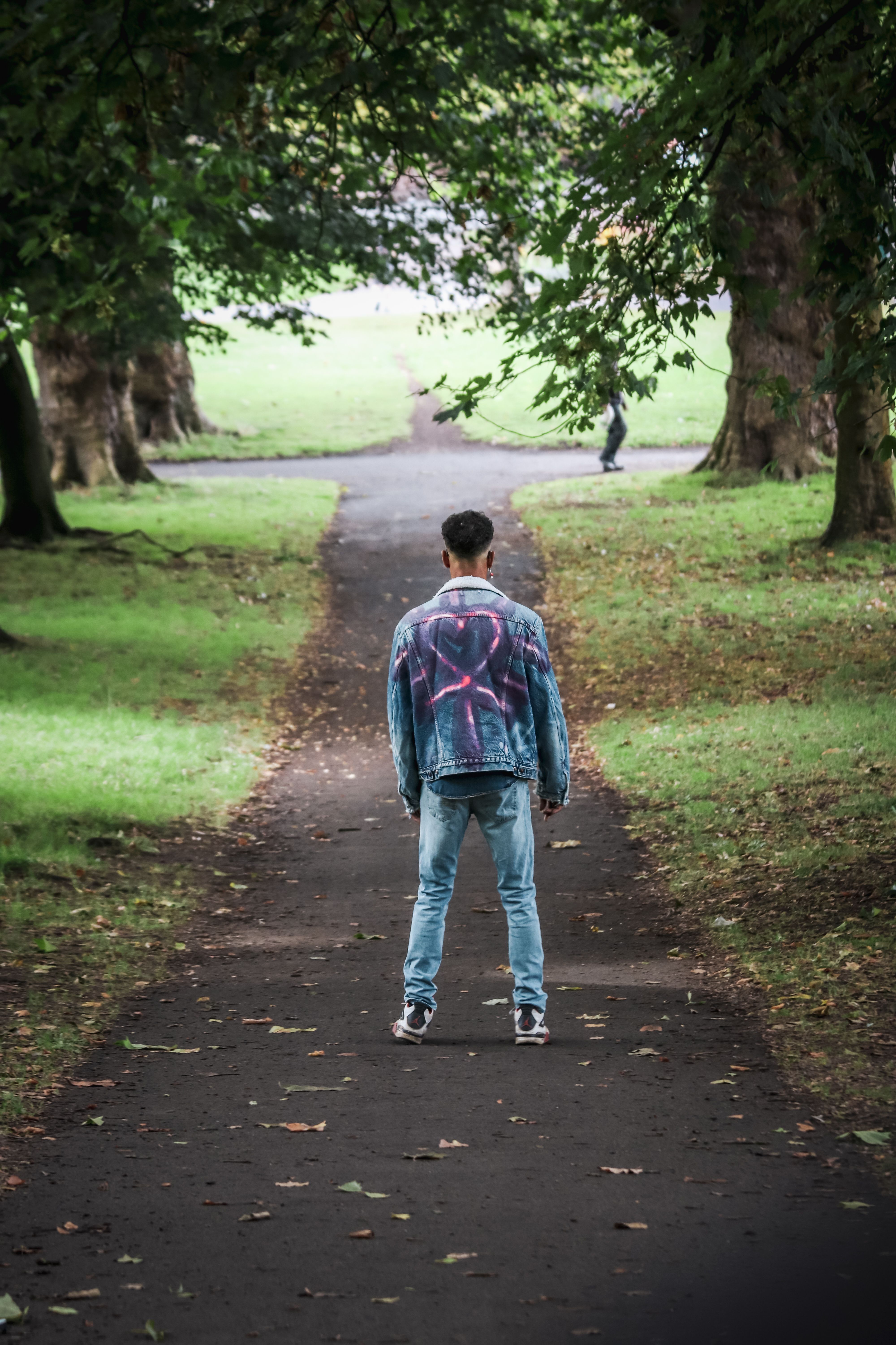 Back view of a young man in a stylish denim jacket strolling along a path in a lush park in Bristol, UK.