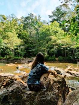 Woman sitting on rocks by a serene river surrounded by lush forest in summer.