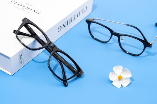 Minimalist display of black eyeglasses on blue background with a white flower and cloth box.