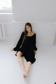 A woman in a black dress poses elegantly while sitting on the floor indoors.