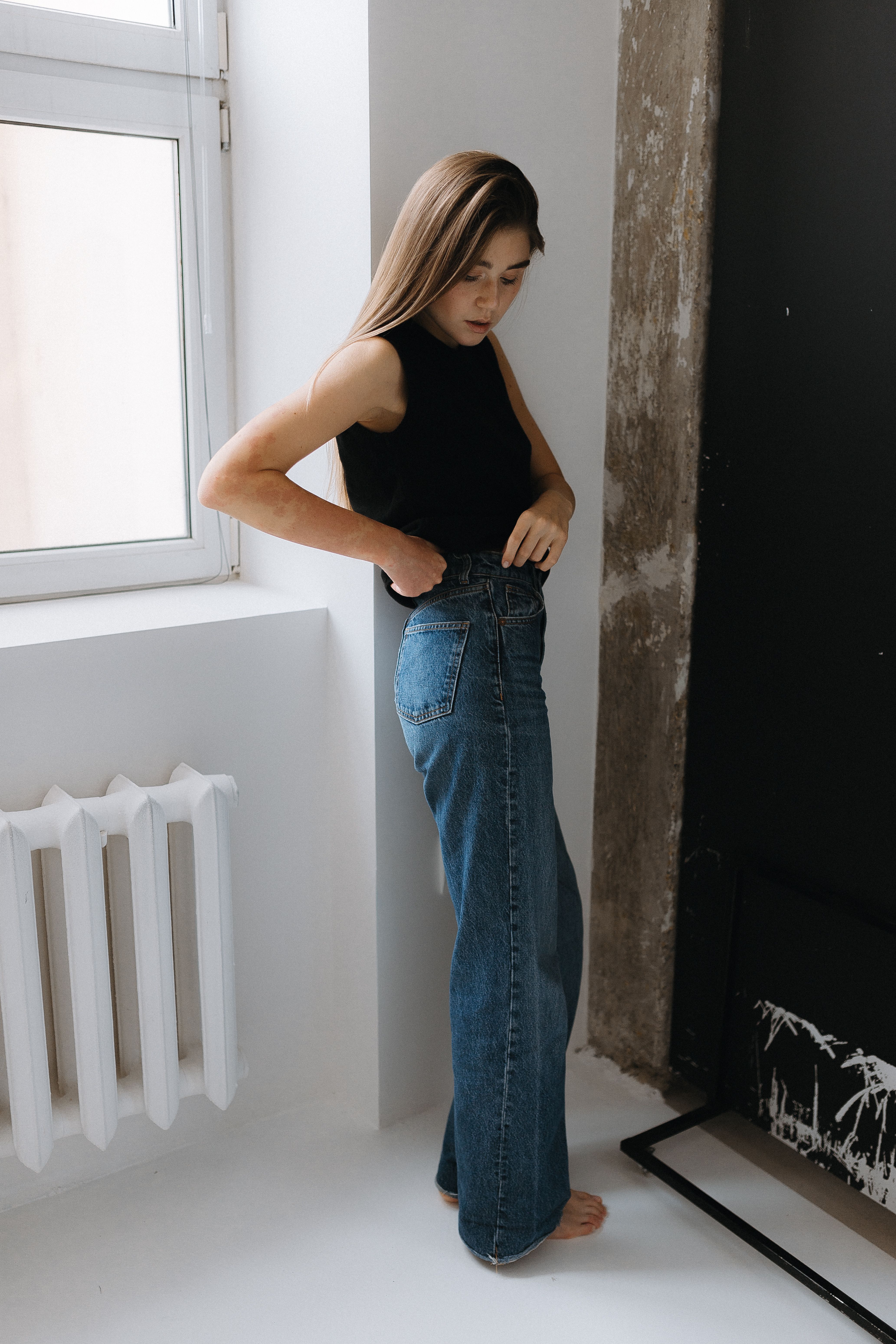 Woman in black top and barrel jeans by a window